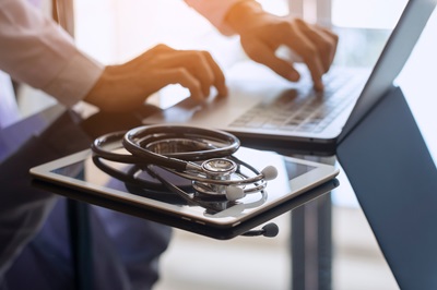 Healthcare provider typing on a laptop with a stethoscope and a tablet next to the laptop