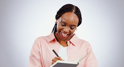 african american woman wearing a pink button down shirt smiling while writing in a notebook