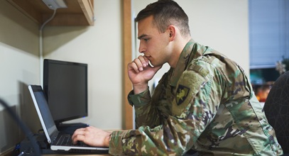 Male military member in uniform sitting at a desk looking at a laptop screen