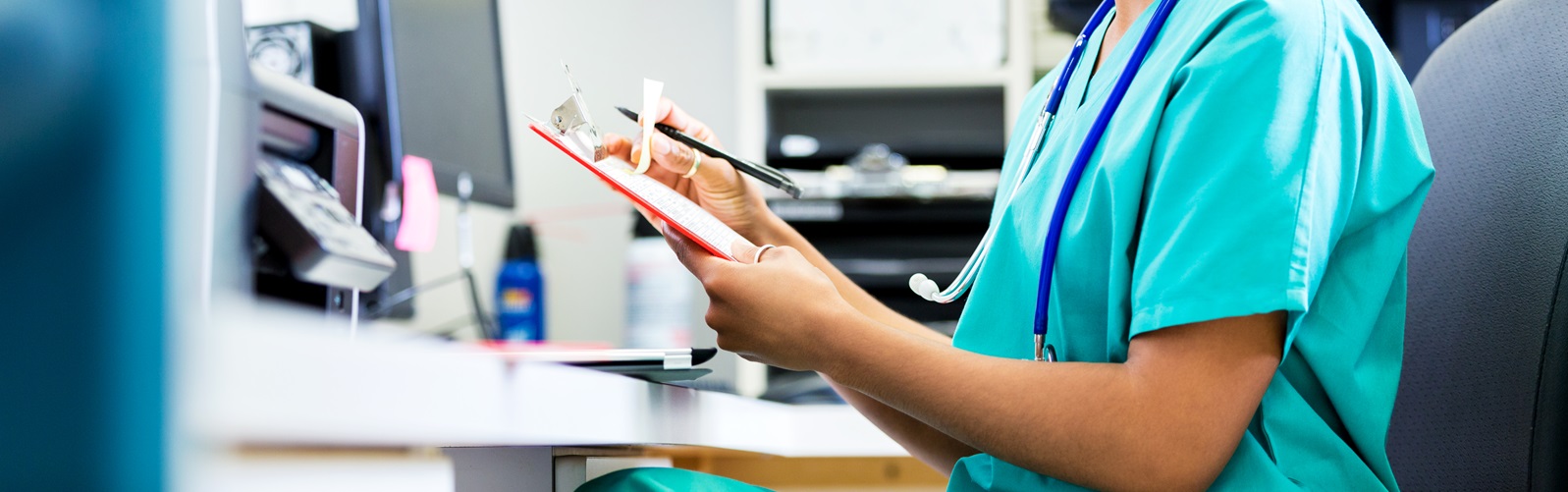 young woman in scrubs sitting at the computer looking at notes on a clipboard