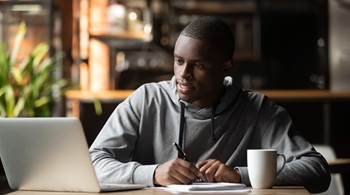 African American man sitting at a desk with a computer, pen and paper, and coffee mug (very self aware)