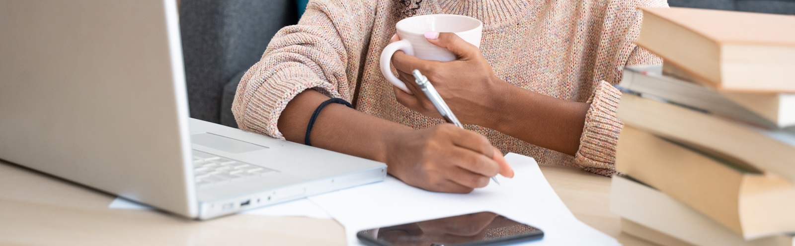 young woman drinking out of a mug, writing on a notepad while looking at her laptop with a pile of books stacked next to her