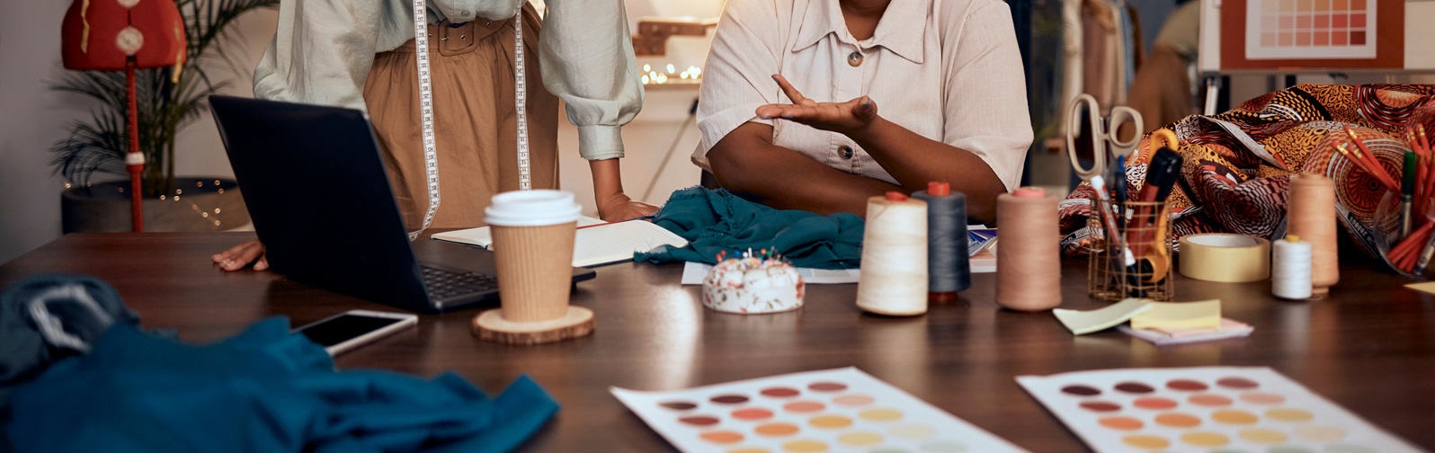 two women discussing their fashion business with a laptop in front of them and many sewing materials across the table