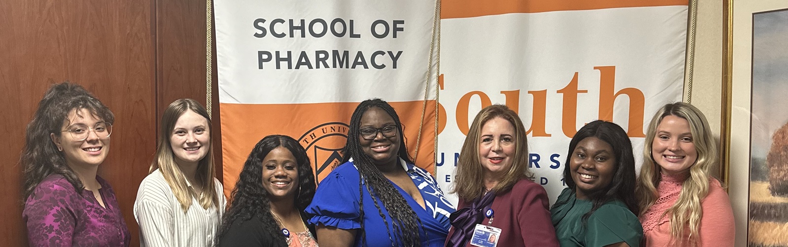 Group of School of Pharmacy women posing in front of the shool's logo