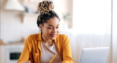 woman with braids in a bun wearing an orange shirt sitting at a desk looking at a laptop