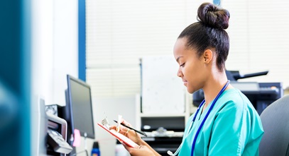 young woman in scrubs sitting at the computer looking at notes on a clipboard